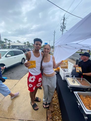 Two people smiling and holding food packets at an outdoor food stall.
