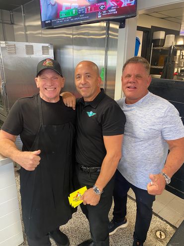 Three men smiling and posing together in a kitchen setting.