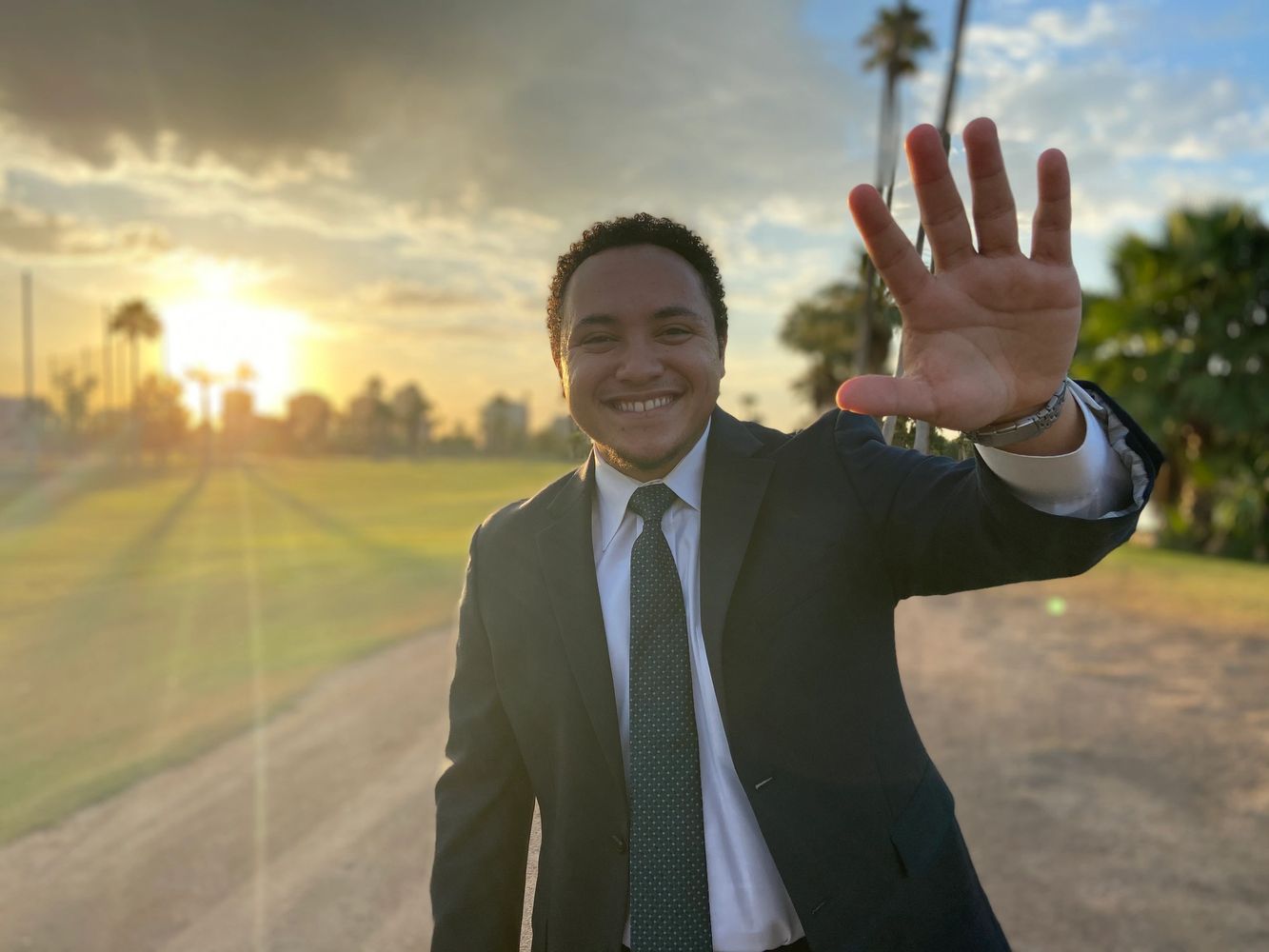 Eric Brock is waving to the camera. A sunrise sits right behind him, peacefully in the distance.