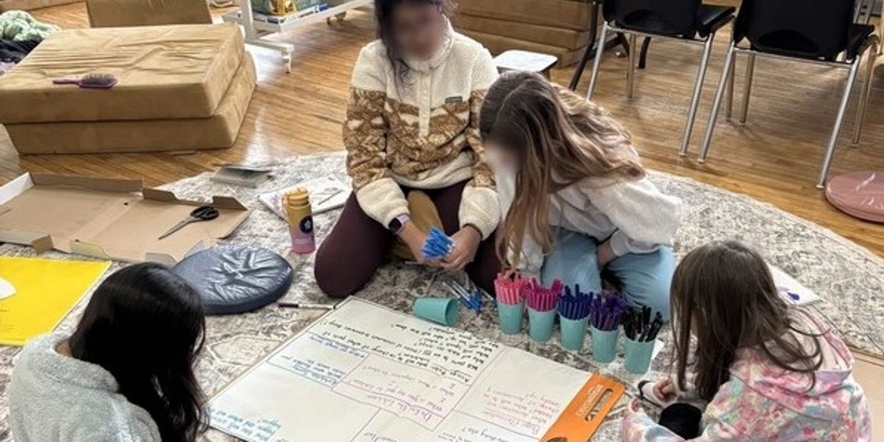 Four children sitting on a rug, writing and organizing colored markers and papers.