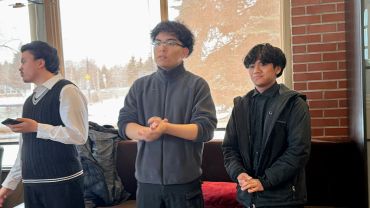 Three young men standing indoors near a window with snowy scenery outside.