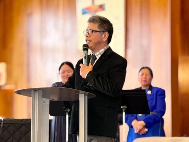Man speaking at a podium in a church setting with two women in the background.
