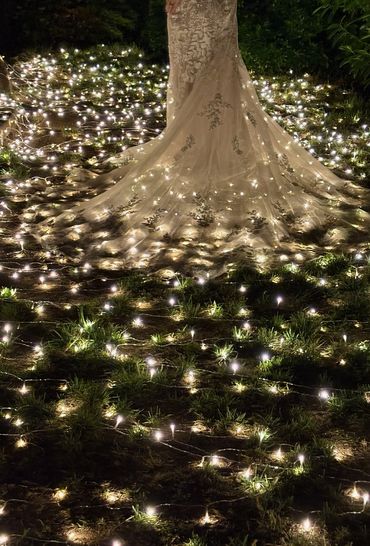 A bride in an elegant gown stands among twinkling fairy lights at night.
