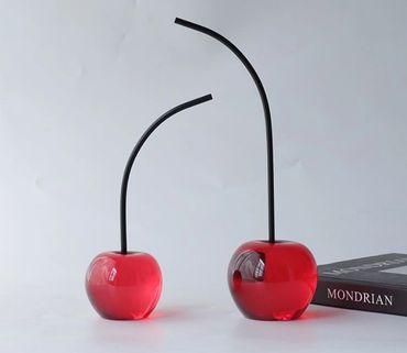 Two decorative glass cherries with long black stems on a white surface next to a book.