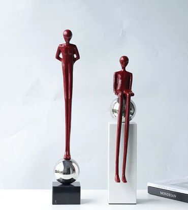 Two elongated red sculptures, one standing and one sitting on reflective spheres.