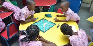 Four children playing Scrabble at a yellow table in school uniforms.