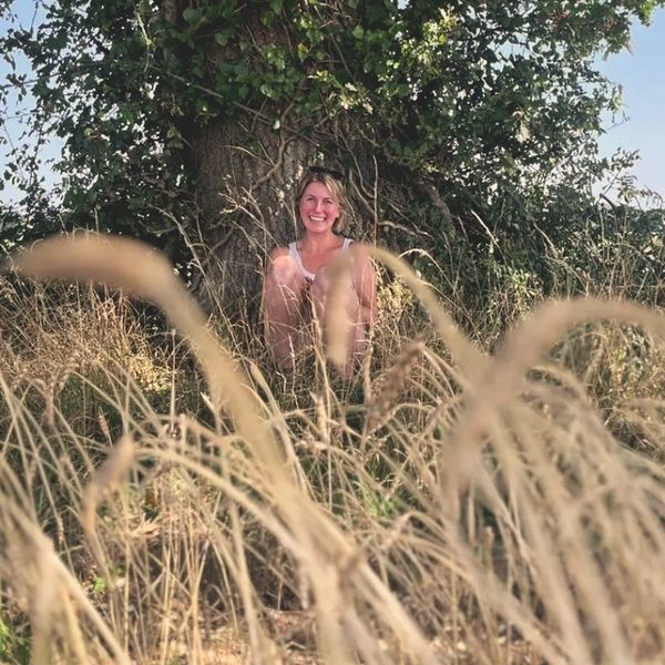 A woman smiles sitting under a tree in tall grass on a sunny day.