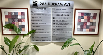 Lobby directory sign listing businesses at 285 Durham Ave with potted plants and framed artwork.
