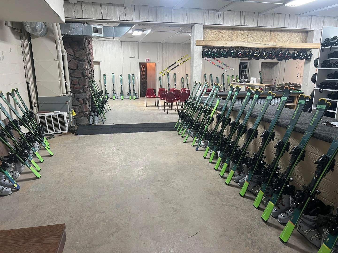 Ski equipment neatly arranged in a rental room with chairs in the back.