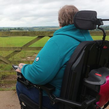 A rear view half body shot of Jayne, sat in her powerchair looking out over green fields in Cornwall