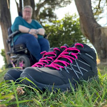 A pair of hiking boots with a blurred image of Jayne sat in her wheelchair in the background