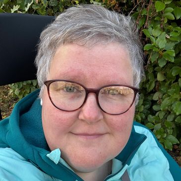 A head shot photo of Jayne smiling. She is wearing glass, has grey hair and a teal green jacket on