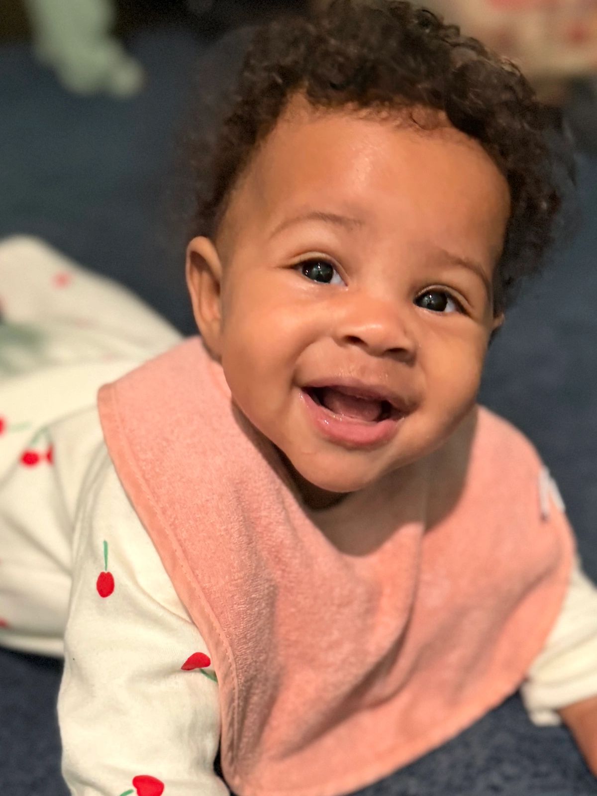Smiling baby with curly hair wearing a pink bib and cherry-patterned outfit.