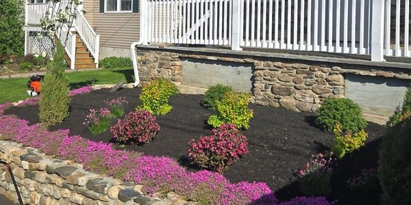 Stone porch house with vibrant flower garden and lawn mower in driveway.