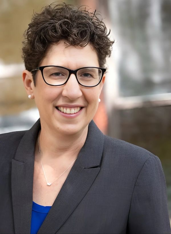 Professional woman with glasses smiling, wearing a dark blazer and blue top.