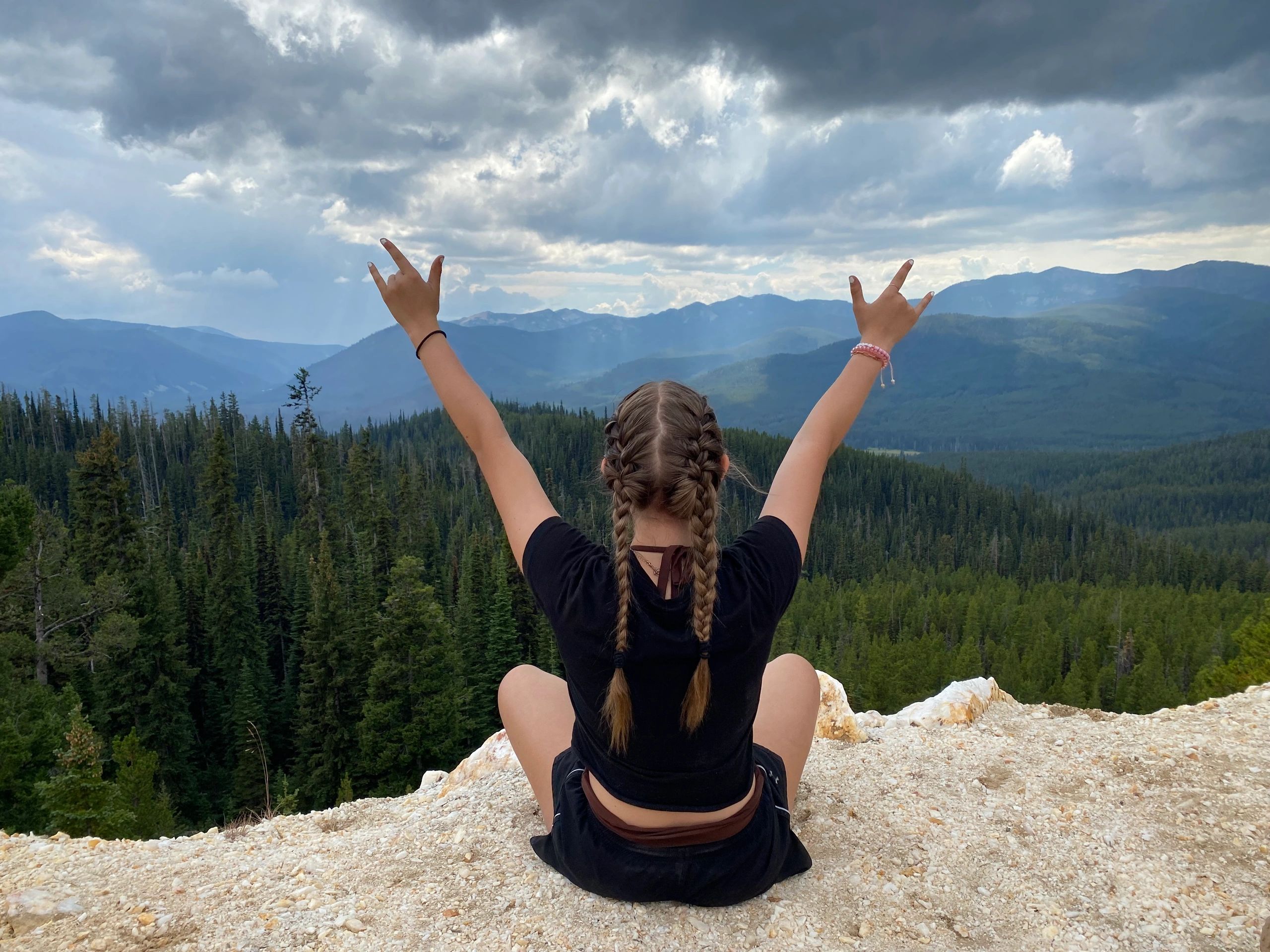 Person with long braids sitting on top of a mountain. Hands raised with the ASL love sign on both.