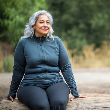 Woman with caramel skin, long gray hair, sitting outside on a log, looking happy
