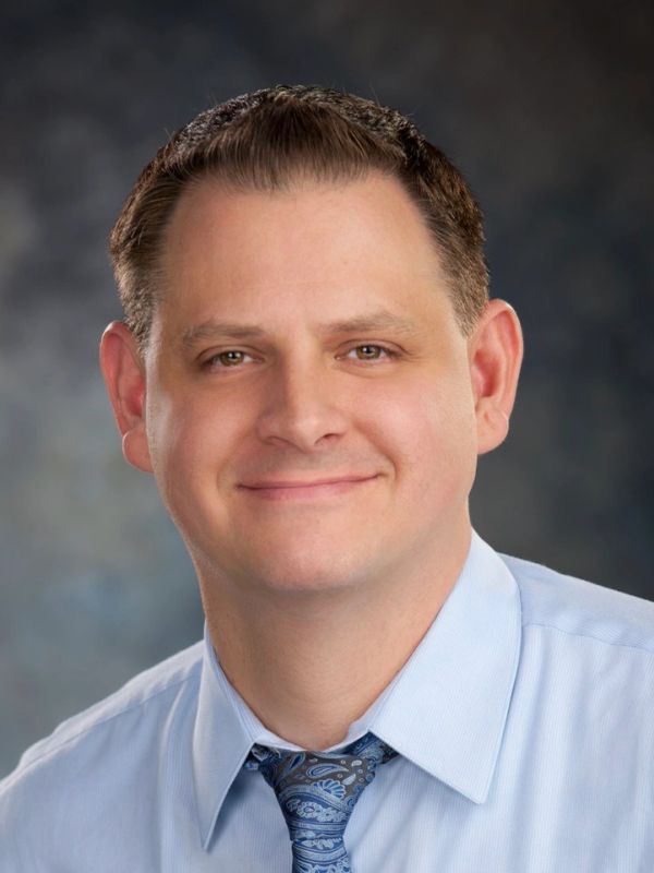 Smiling man in a blue shirt and patterned tie against a blurred background.