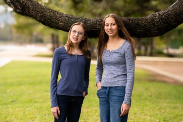 Two teenage girls standing outdoors under a large tree branch, smiling at the camera.