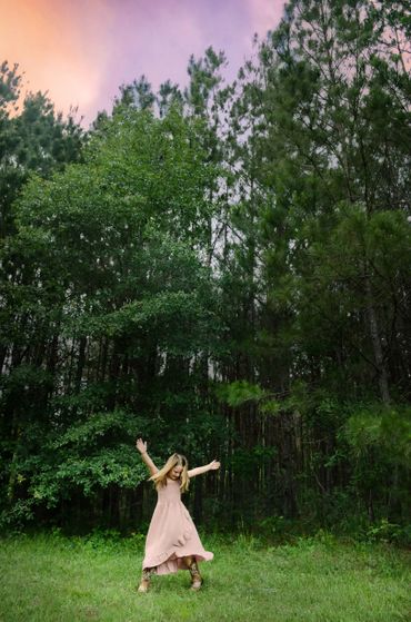 Young girl dances joyfully in a grassy clearing near tall trees at sunset.