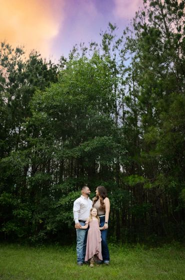 A family of three standing in front of tall trees during sunset.