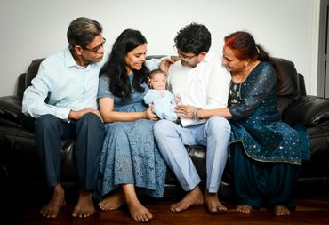 A joyful family of four generations gathered around a newborn baby on a couch.