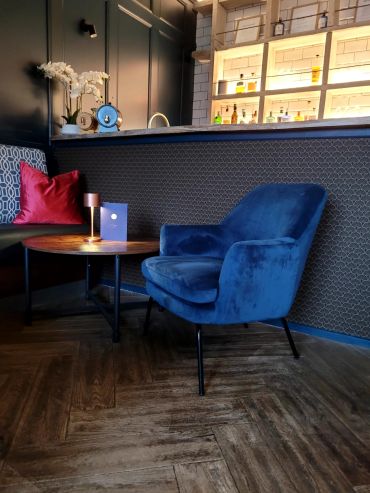 Cozy seating area with a blue velvet chair and illuminated bar shelves.