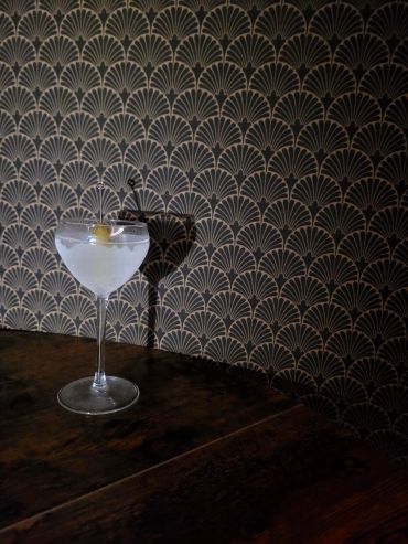 A cocktail glass with olive against patterned wallpaper.