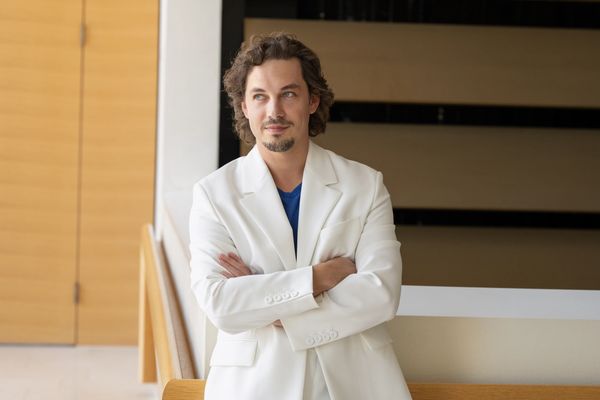 Man in a white suit leaning on a railing, looking thoughtfully to the side.