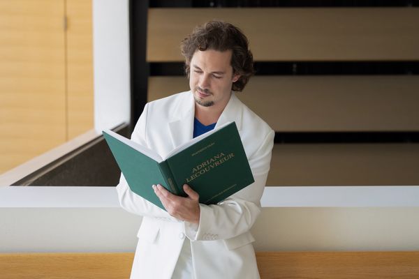 Man in white suit reading a large green book indoors.