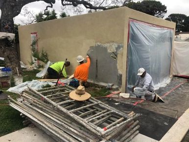 Three workers plastering a small building under cloudy skies.