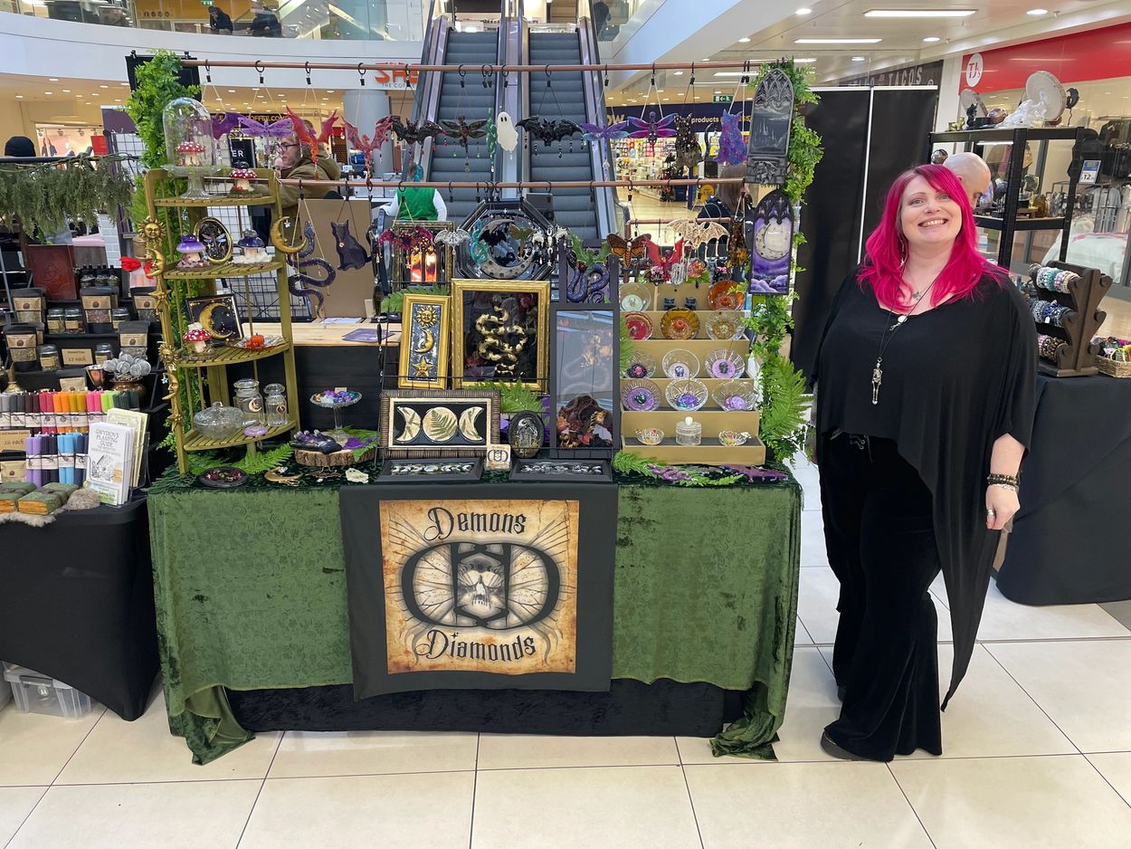 A vendor with bright pink hair stands beside her mystical-themed craft booth in a mall.