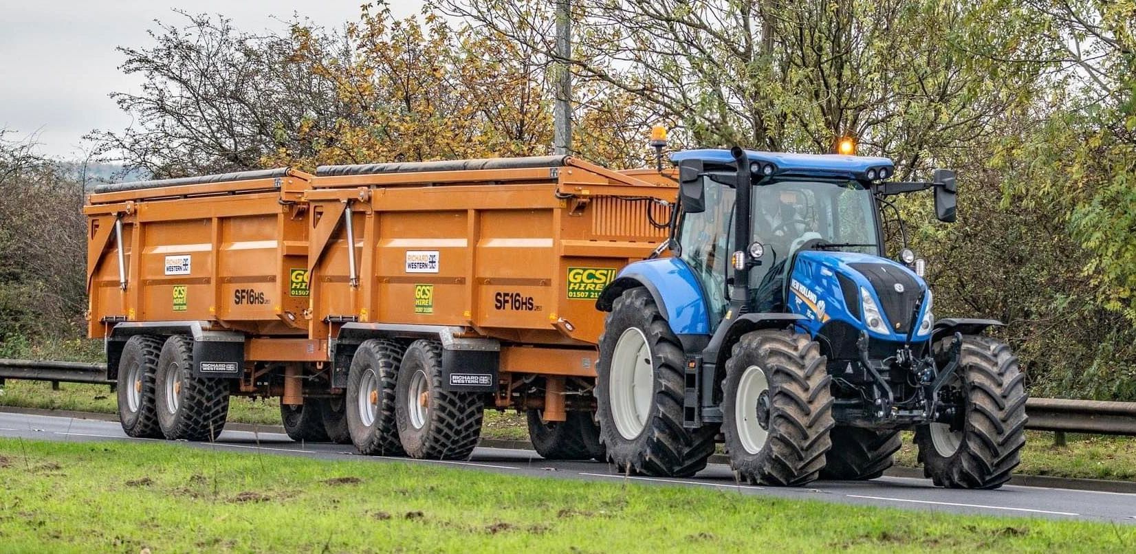 Blue tractor pulling two large orange trailers on a road.