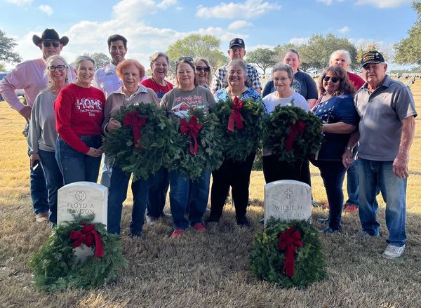 Group of people honoring graves with wreaths in a sunny cemetery.