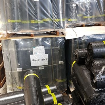 Rolls of black material labeled 'Made in Egypt' stacked on pallets in a warehouse.