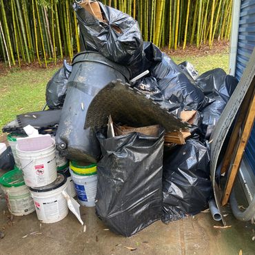 Pile of black garbage bags, buckets, and trash on a concrete floor near bamboo plants.