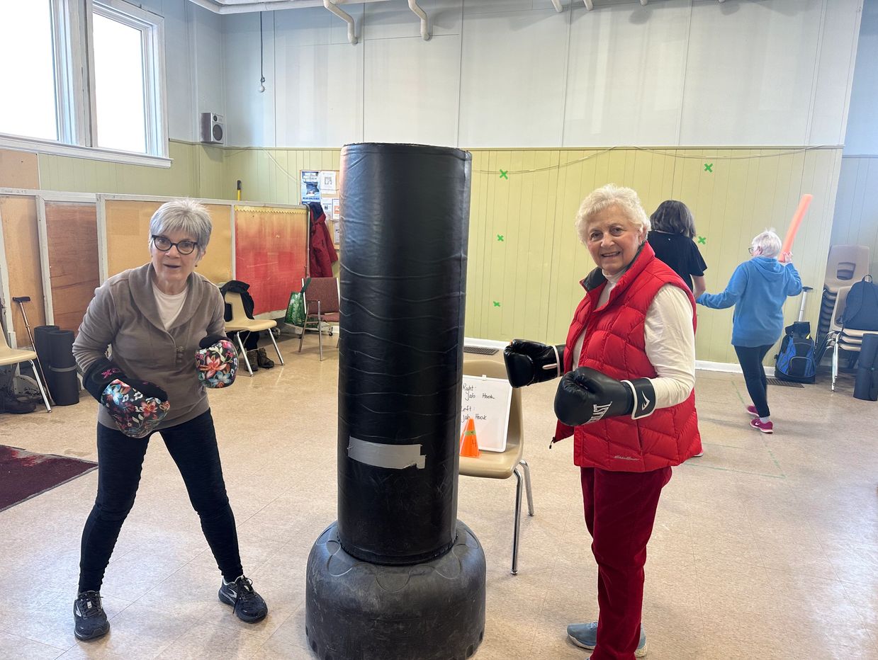 Two elderly women with boxing gloves pose by a punching bag indoors.