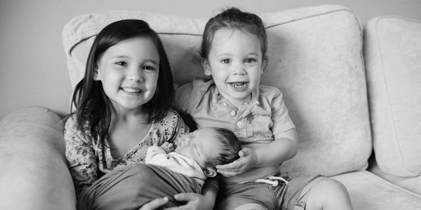 Three siblings sitting on a couch, smiling with the baby asleep.