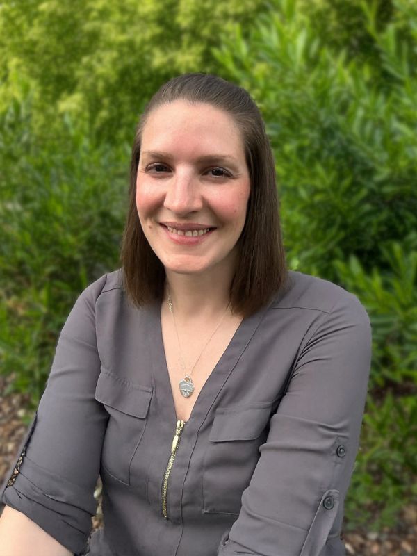 Smiling woman in gray top outdoors with greenery background.