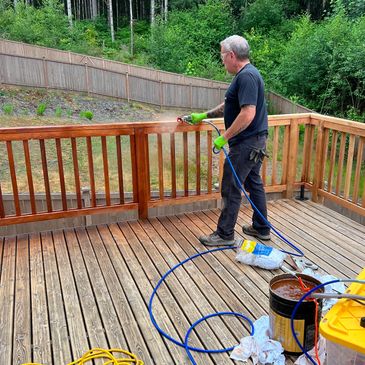 Man spray-painting a wooden deck railing with a sprayer outdoors.