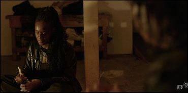 A woman in a leather jacket sitting and stirring something in a jar in a dimly lit room.