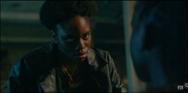 A serious woman wearing a red pendant necklace looks intently in dim lighting.