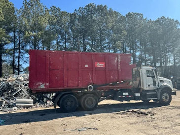 White truck carrying a large red container in a scrapyard.