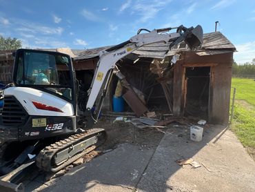Excavator demolishing an old wooden shed under a clear sky.