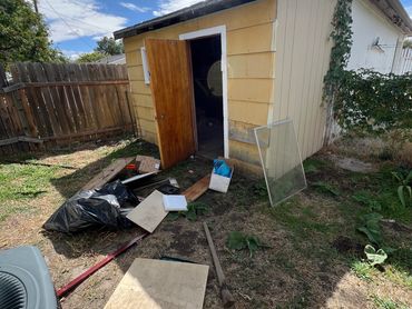 A yellow shed with an open door and scattered debris outside on a sunny day.