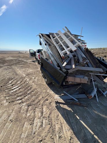 Truck with open trailer carrying wooden debris on a dirt road.