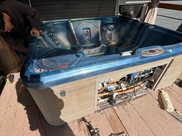 Person repairing a blue hot tub on a wooden deck under sunlight.