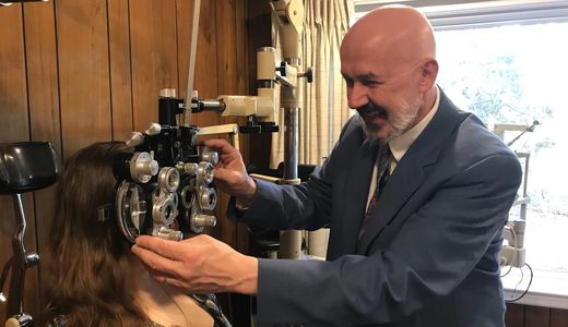 A smiling Dr. Kofos places a phoropter in front of a patient's eyes.