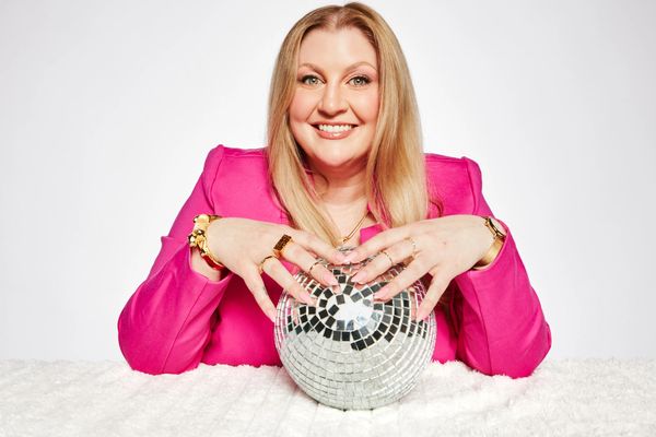 Smiling woman in a pink blazer holding a disco ball on a white surface.