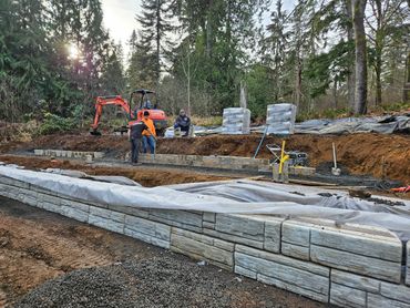 Workers building a stone retaining wall in a forested area with construction equipment.